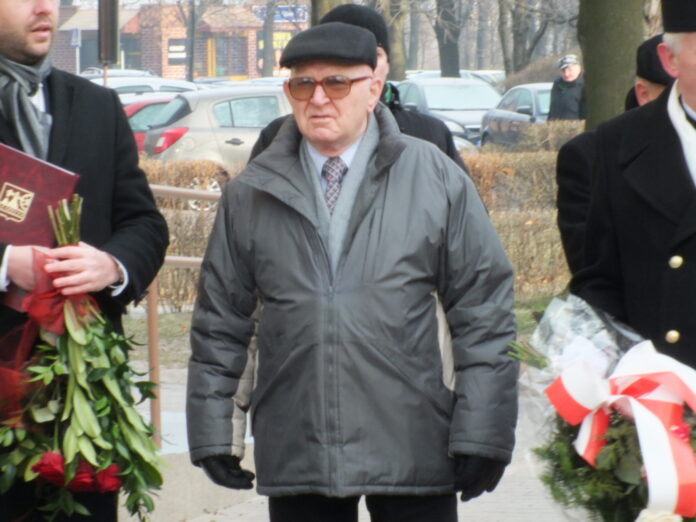 Na zdjęciu jest Jan Ludwiczak, ubrany w kurtkę i czapkę. Obok niego stoją osoby z wiązankami kwiatów