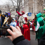 Orszak Trzech Króli wraca do Katowic Zdjęcie przedstawia trzech króli na koniach. Na pierwszym planie widać telefon komórkowy, którym ktoś robi zdjęcie królom. W tle kilkadziesiąt osób obserwujących króli i budynek katowickiej katedry.