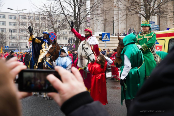 Zdjęcie przedstawia trzech króli na koniach. Na pierwszym planie widać telefon komórkowy, którym ktoś robi zdjęcie królom. W tle kilkadziesiąt osób obserwujących króli i budynek katowickiej katedry.