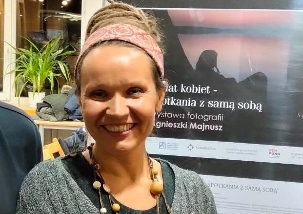 na zdjęciu widać autorkę zdjęć Agnieszkę Majnusz, która uśmiechnięta stoi obok plakatu promującego wystawę