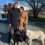 Niepełnosprawność i sukces: inspirująca rozmowa z Bartoszem Radomskim i Martyną Zadorożną Mężczyzna po lewej stronie ma na sobie czarny płaszcz, a kobieta po prawej stronie ma na sobie brązowy płaszcz. Oboje trzymają smycze dwóch psów: czarnego i białego. W tle widać duży statek z masztami oraz drzewo bez liści. Na statku powiewa polska flaga. Cała scena ma miejsce w słoneczny dzień na wybrukowanej ścieżce obok trawnika.