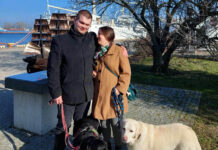 Niepełnosprawność i sukces: inspirująca rozmowa z Bartoszem Radomskim i Martyną Zadorożną Mężczyzna po lewej stronie ma na sobie czarny płaszcz, a kobieta po prawej stronie ma na sobie brązowy płaszcz. Oboje trzymają smycze dwóch psów: czarnego i białego. W tle widać duży statek z masztami oraz drzewo bez liści. Na statku powiewa polska flaga. Cała scena ma miejsce w słoneczny dzień na wybrukowanej ścieżce obok trawnika.