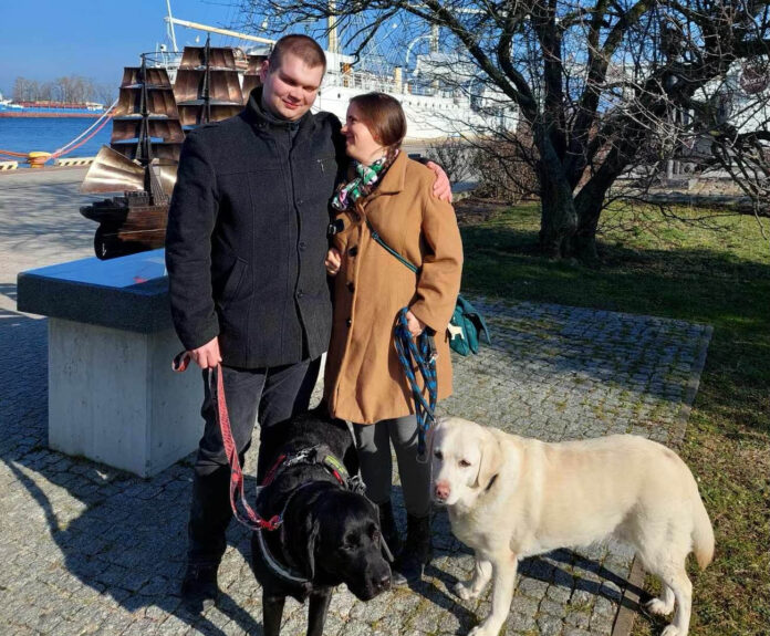 IMG_3826 kopia Mężczyzna po lewej stronie ma na sobie czarny płaszcz, a kobieta po prawej stronie ma na sobie brązowy płaszcz. Oboje trzymają smycze dwóch psów: czarnego i białego. W tle widać duży statek z masztami oraz drzewo bez liści. Na statku powiewa polska flaga. Cała scena ma miejsce w słoneczny dzień na wybrukowanej ścieżce obok trawnika.