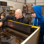 Szósta edycja programu „Katowice Miastem Fachowców” Mężczyzna pokazuje funkcjonowanie maszyny chłopakowi ubranemu w bluzę z kapturem