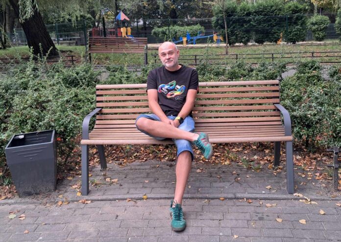 Bartosz Danowski siedzi na łąwce, w tle widać plac zabaw