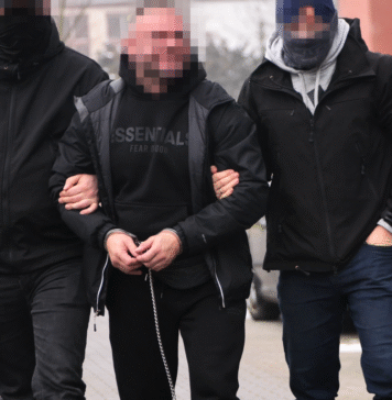 Maczety, siekiery i kokaina. Śląska policja uderzyła w brutalny gang pseudokibiców Torcidy i GieKSy Dwóch nieumundurowanych policjantów prowadzi zakutego w kajdanki mężczyznę z zakrytą twarzą; funkcjonariusze mają na rękawach opaski z napisem „Policja”