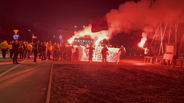 Grupa kibiców z transparentem „Gloria Victis” i odpalonymi racami podczas nocnego przemarszu w Katowicach, czerwony dym nad pomnikiem.